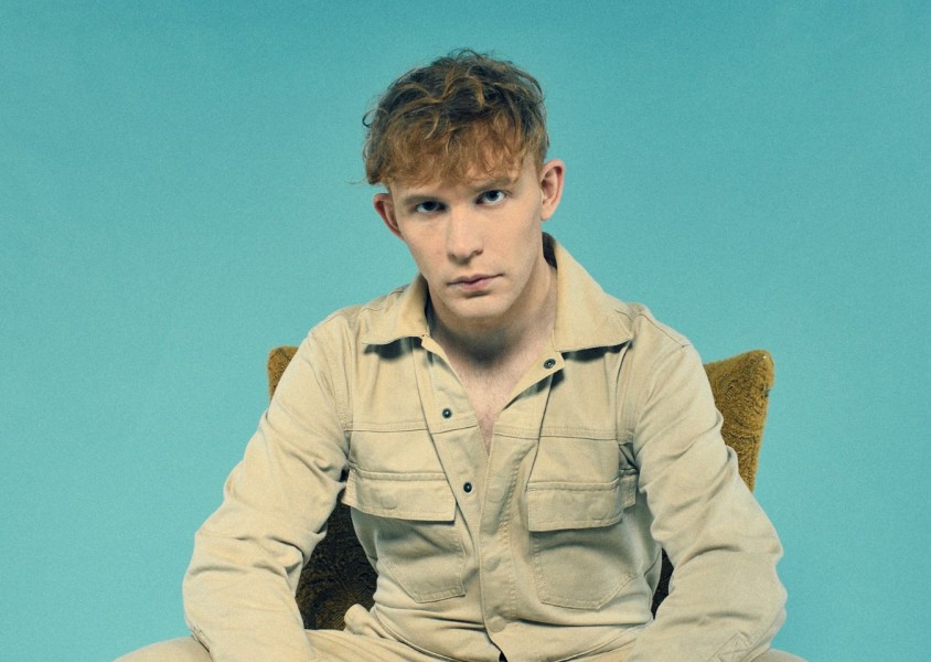 A guy sat looking towards the camera in front of a blue backdropx